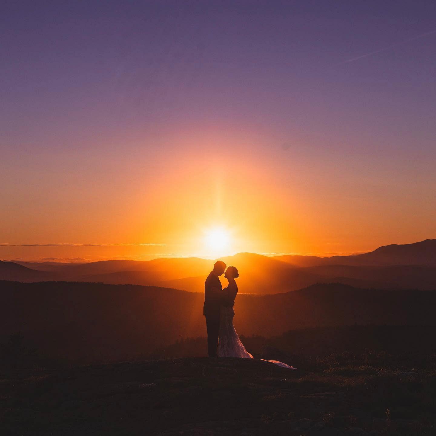 a couple standing looking at each other on a hilly terrain while the sun rises from behind them