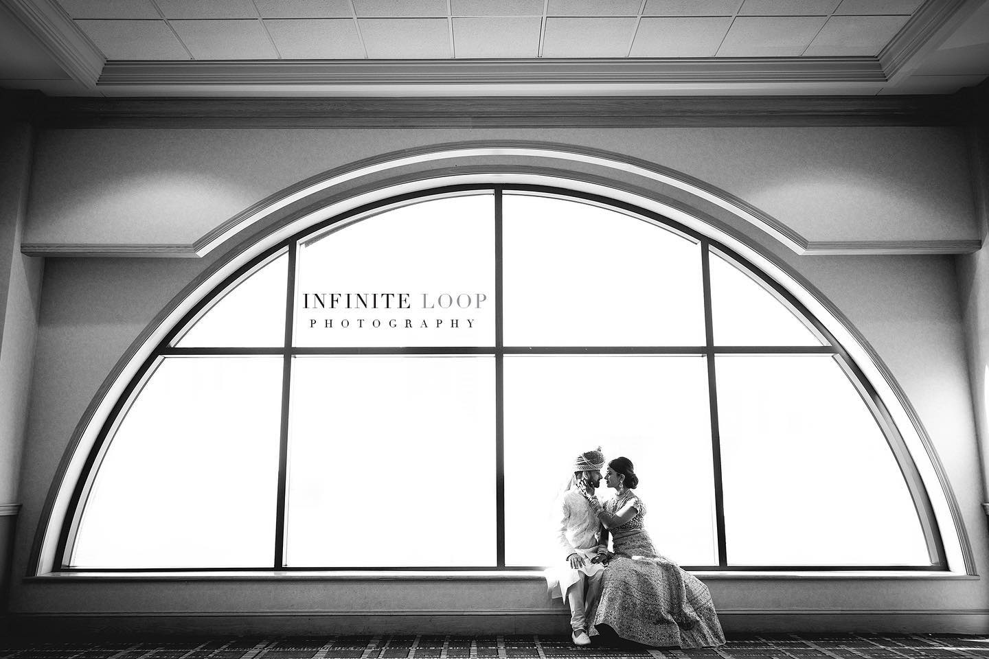 Black and white photo of a bride and groom dressed in traditional clothes pose in front of a semi-circle shaped window