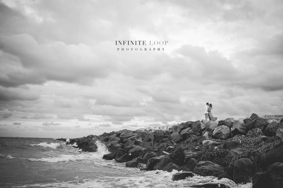 Black and white photo of a couple kissing on top of a cliff as the waves crash on the rocky shore 