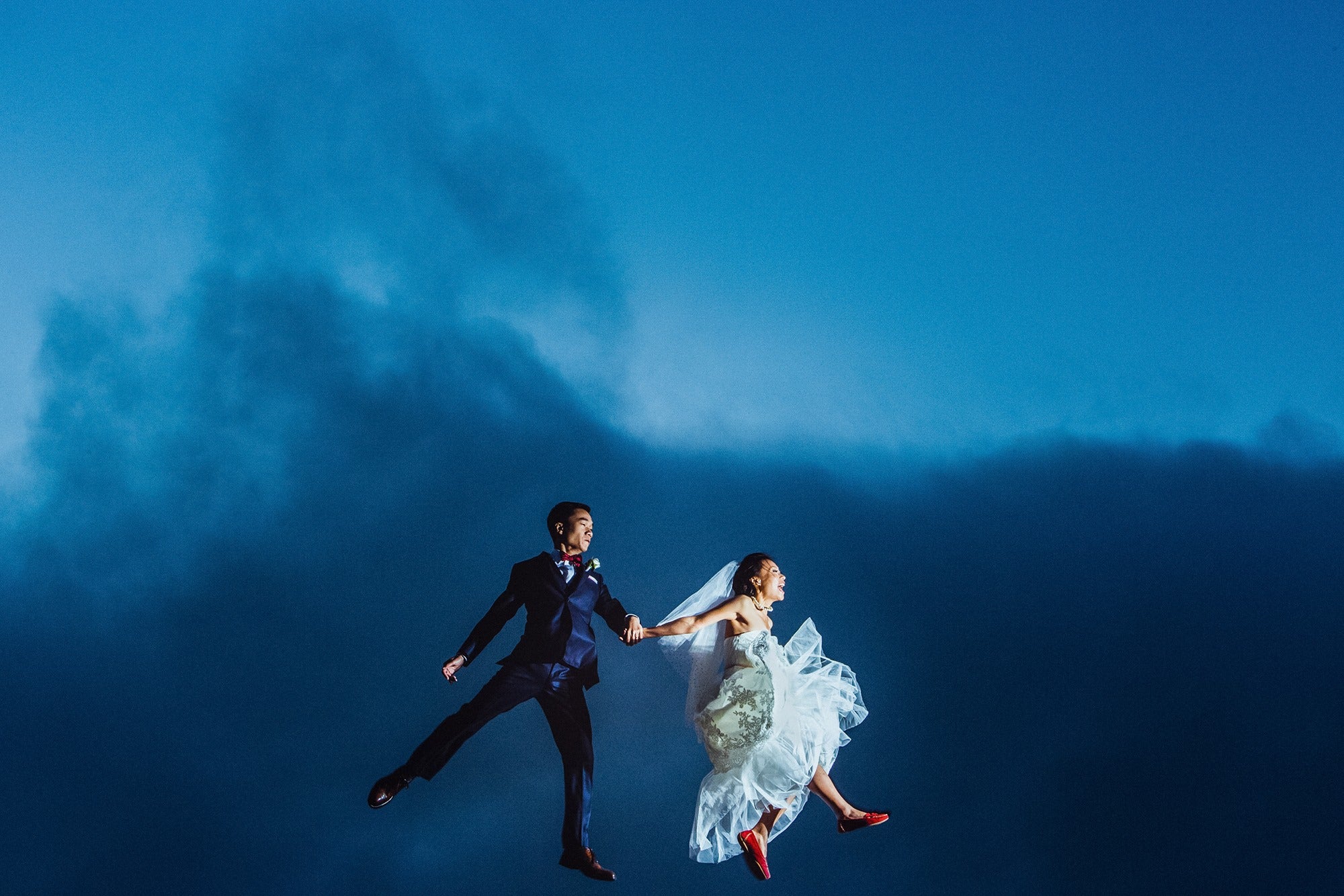 Bride and groom holding hands during a jump shot against a blue sky