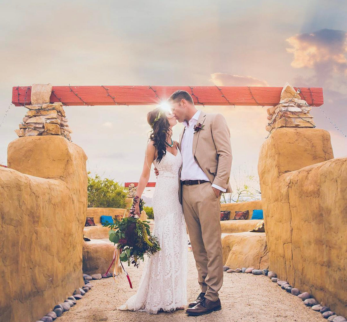 a recently married couple standing between a stone gate and kisses while the sun shines between them