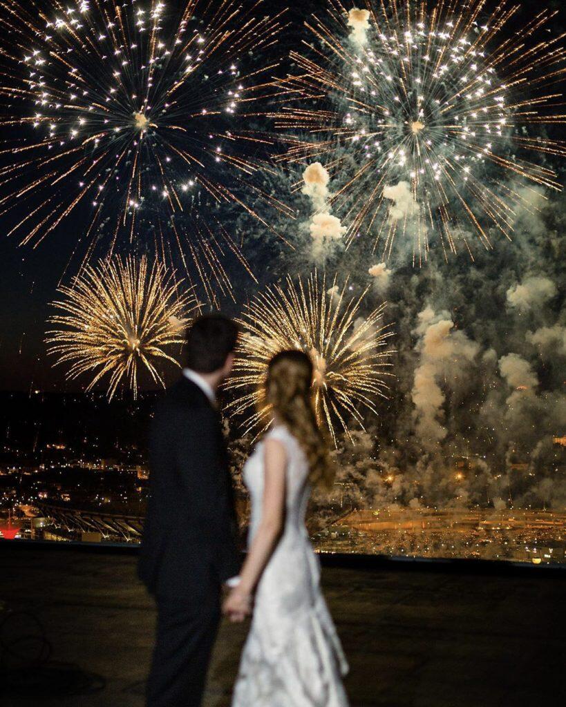 a newlywed couple holding hands and watching a fireworks display