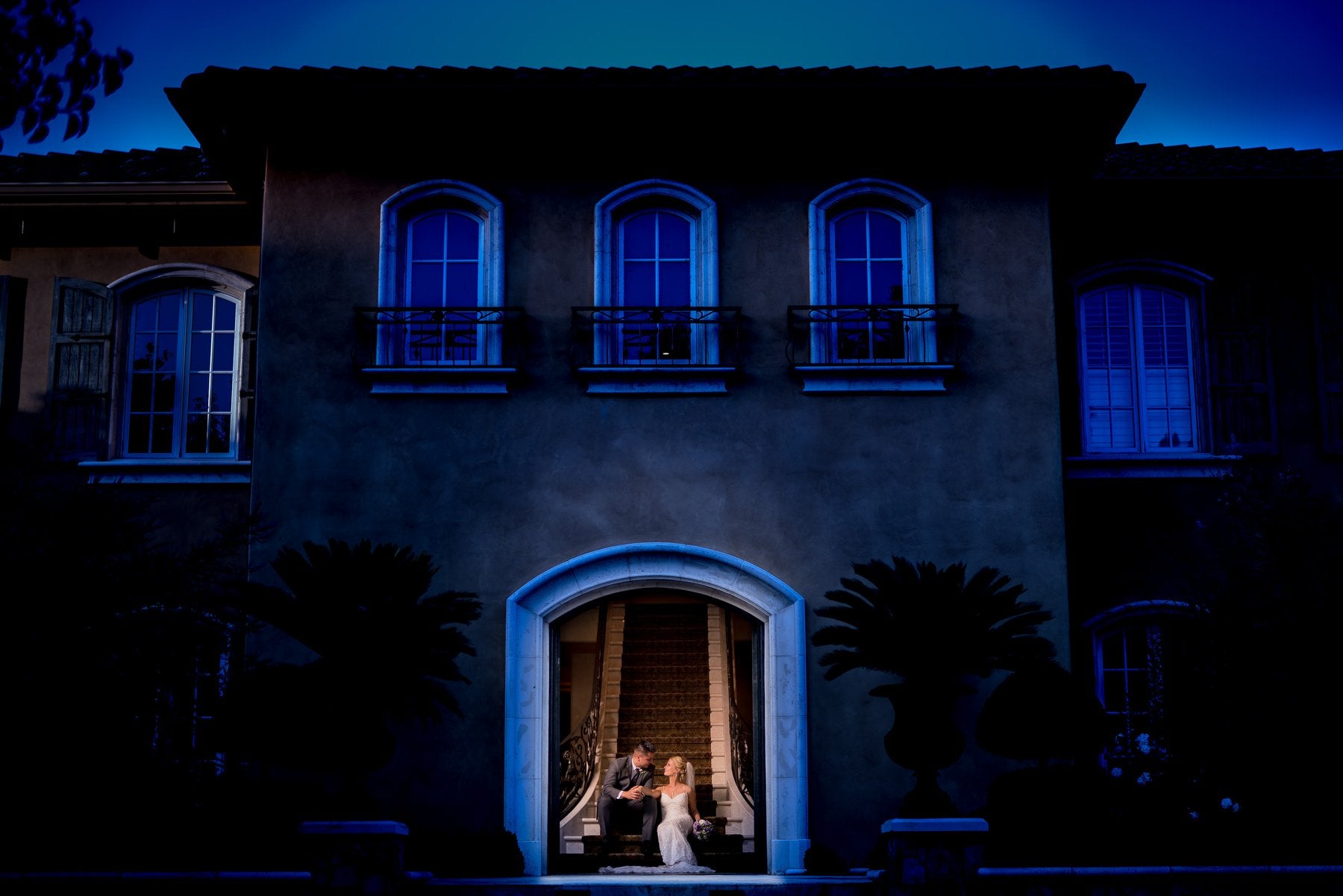 Blue tinted dark and moody wedding photo of a bride and groom sitting in at the front of a stairway of a place