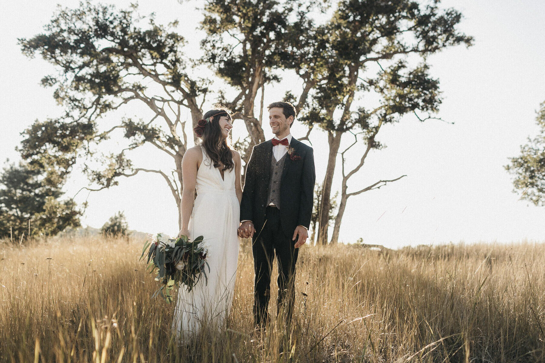 a wedding couple standing in a grass field looking at each other smiling with the bridal bouquet in hand