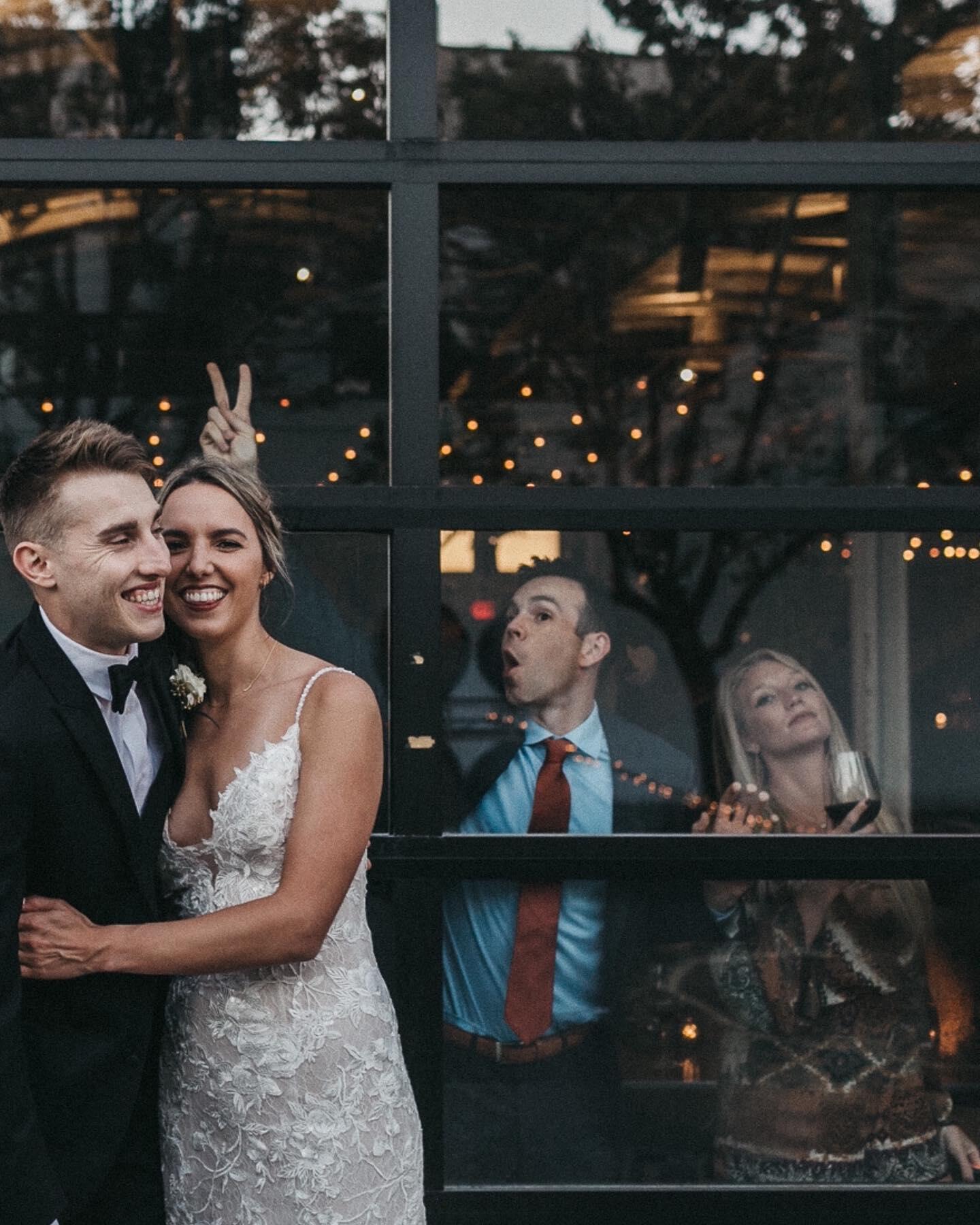 a recently married couple posing in front of a glass panel while their friends make funny gestures from behind