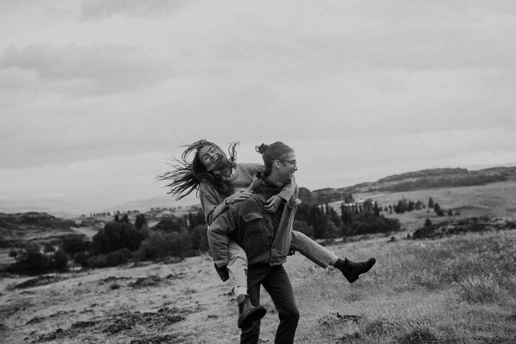 a couple on a grass field where the groom is giving a piggy back ride to the bride