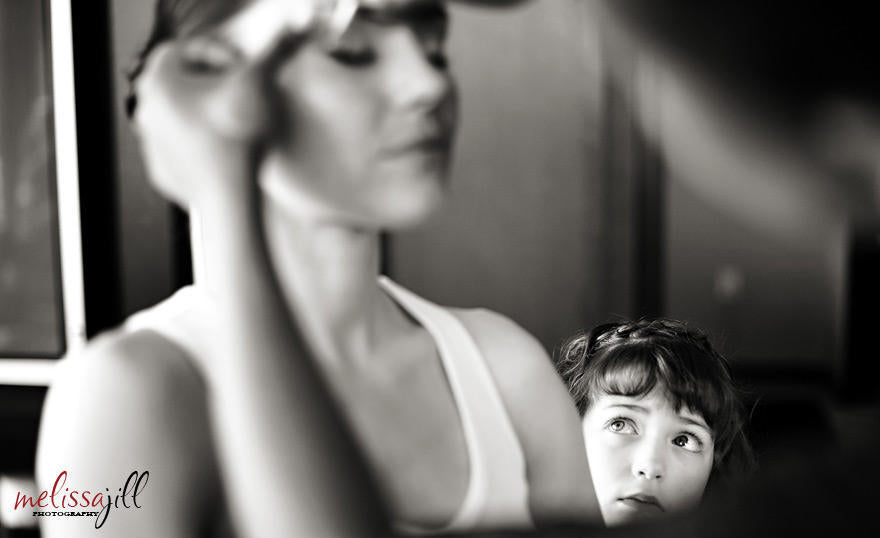 A black and white wedding photography image of the bride getting her makeup done, while a young girl watches from the side.