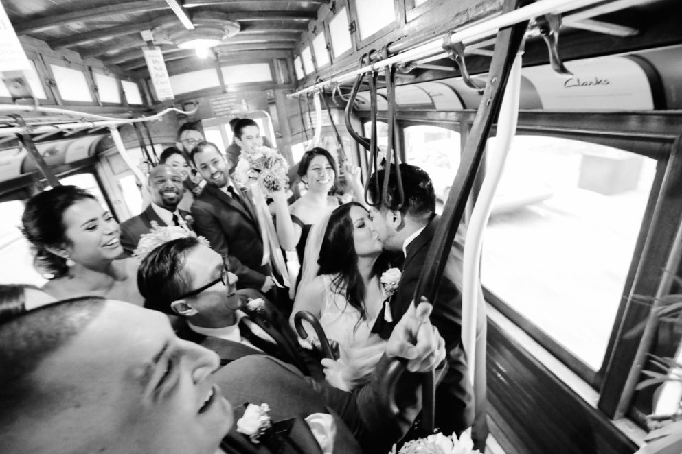 Bride and groom share a kiss on a subway as the guests surrounding them cheer with joy