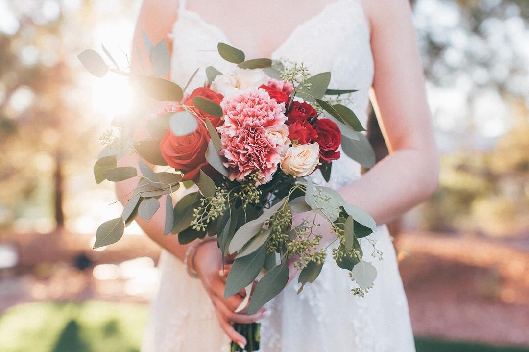 Close up shot of a bride’s hand holding a bouquet with red, pink, and white flowers.