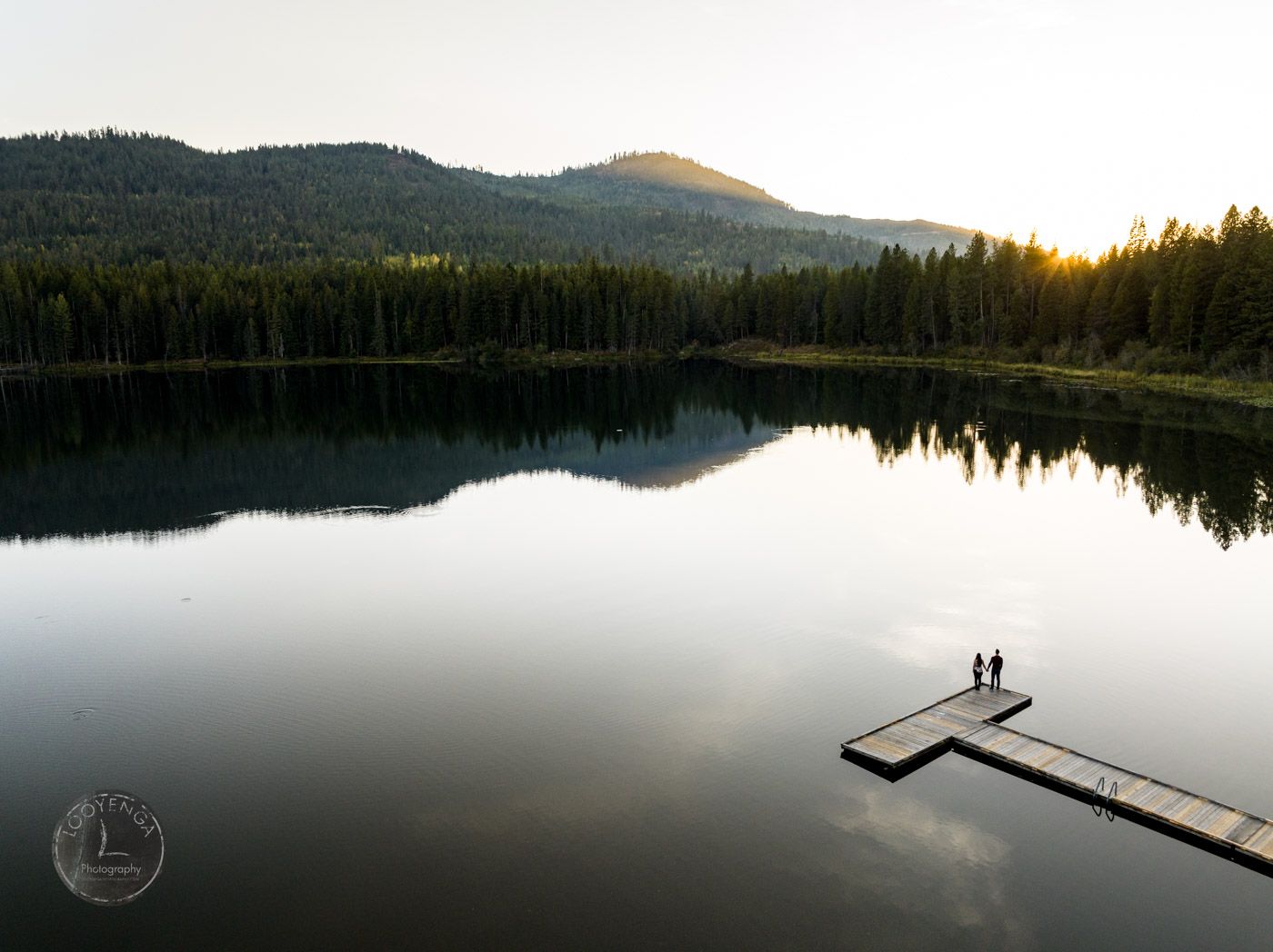 a couple standing on a pier by a lake surrounded by forest