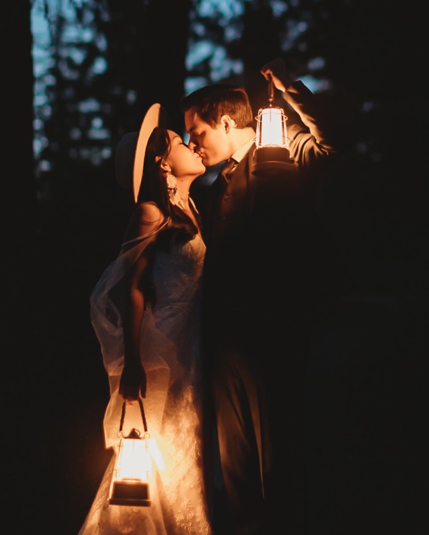 a couple kissing standing in the woods holding lanterns in their hand