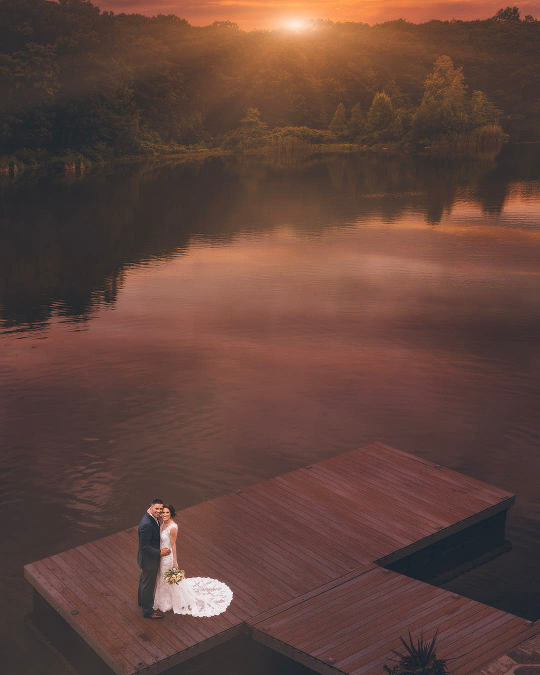 a couple standing on a pier in their wedding dress by a big lake during sunset