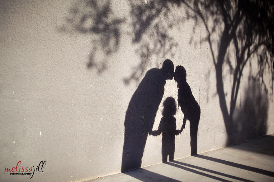 A family photography image of the couple kissing with their child in the middle as a shadow on a wall outdoors.