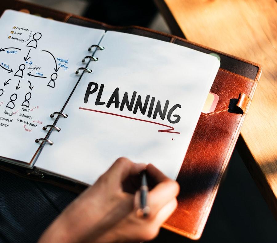 A woman's hand holding a pen over a notebook with photography workflow diagram & the word “planning” with a red underline.