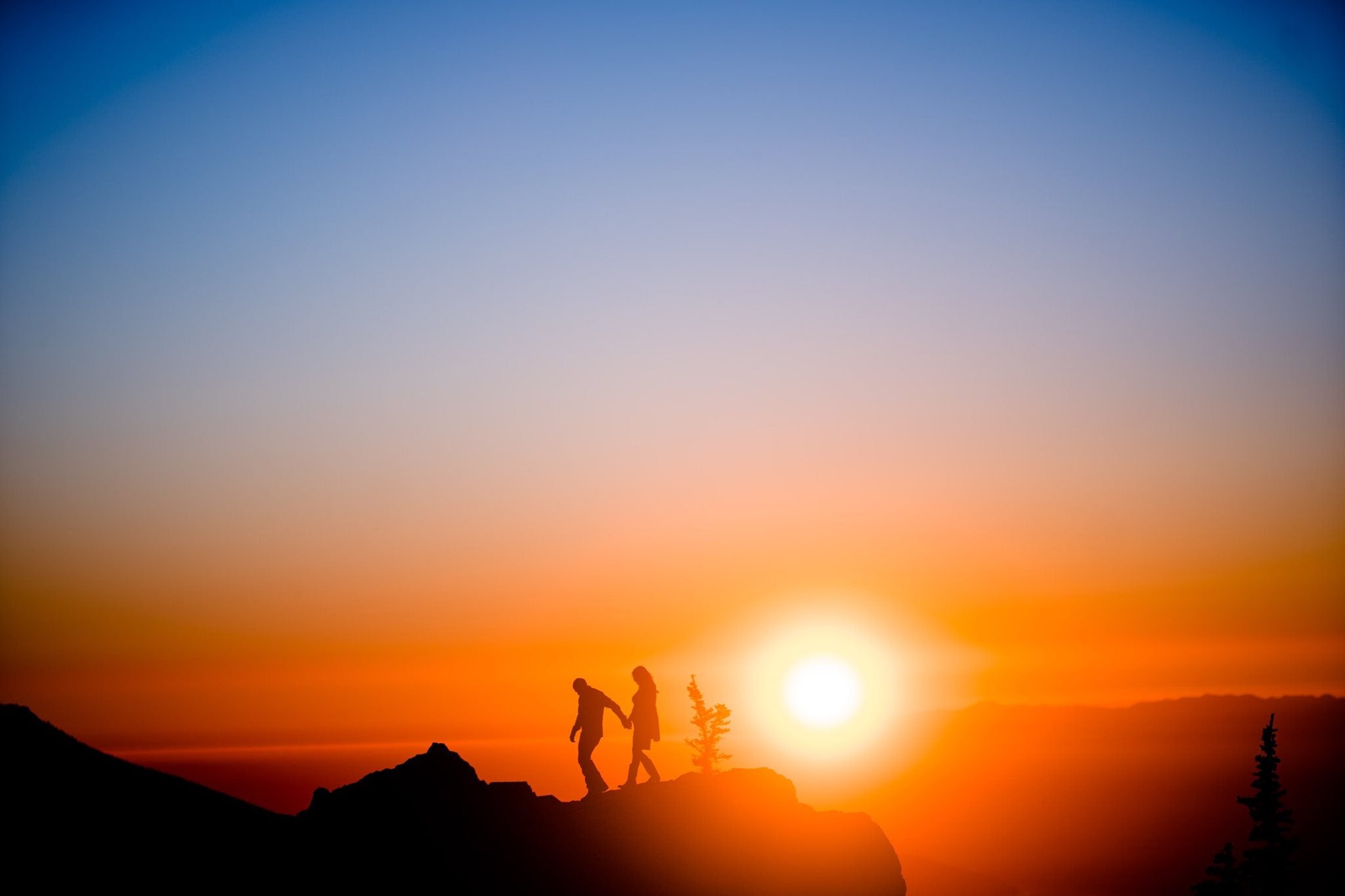 Silhouette of a couple walking holding hands on top of a cliff as the sun sets behind them