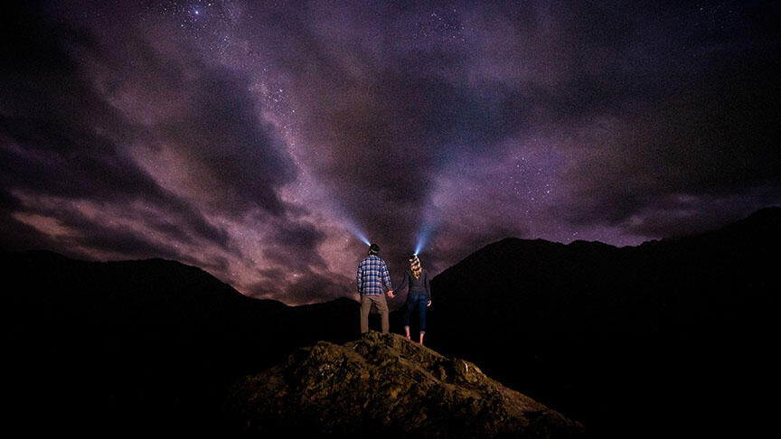 couple standing on a rock during a starry night wearing headlamps