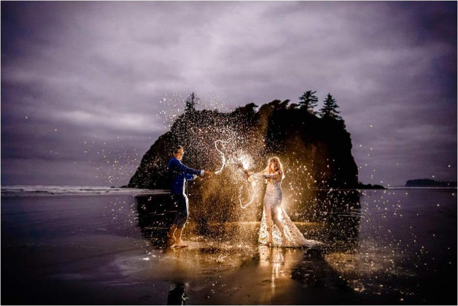 couple popping a champagne bottle and spewing champagne everywhere near a rock in the ocean wearing a wedding dress and tux