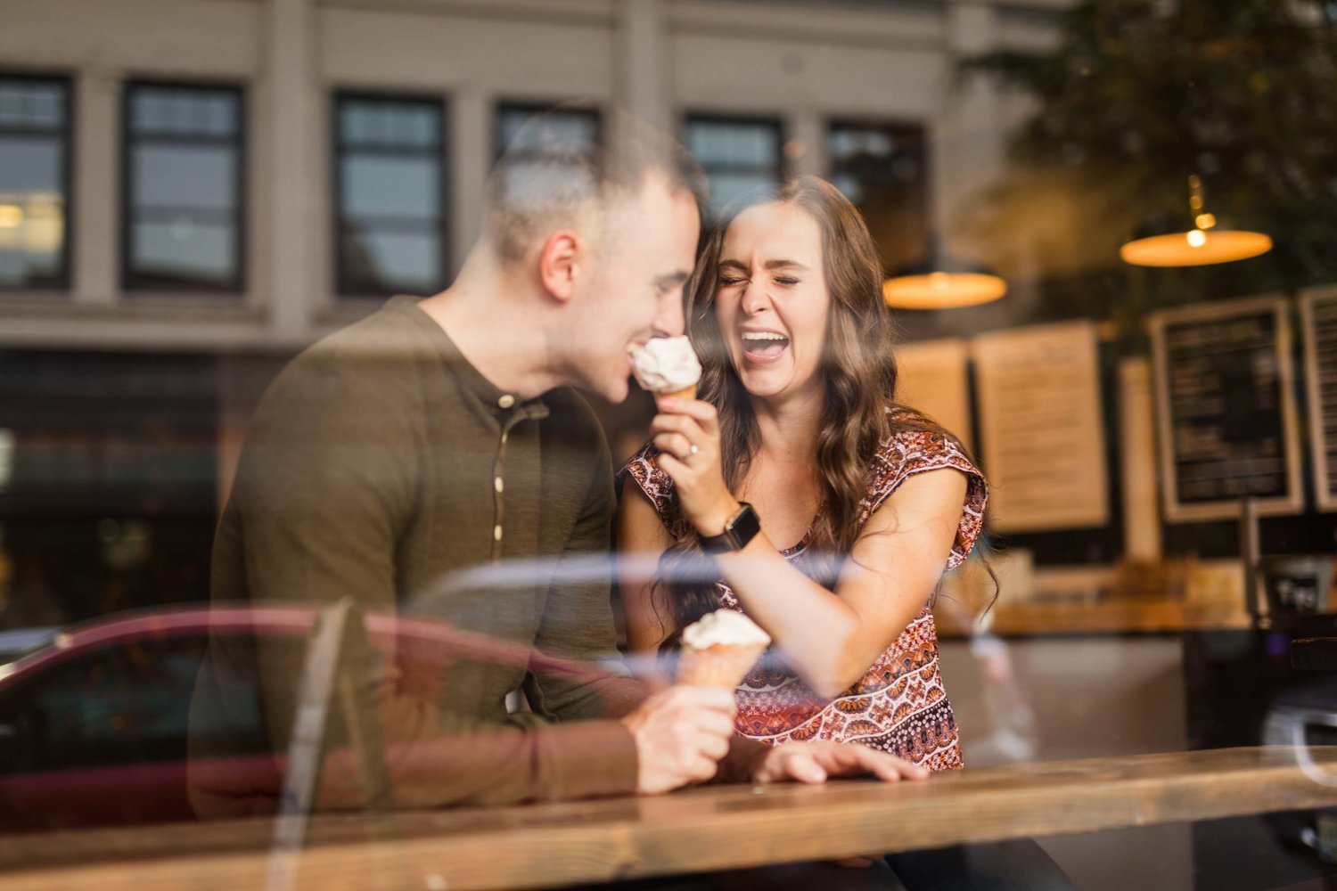 A portrait of a couple through the window of a café as they laugh sharing ice creams