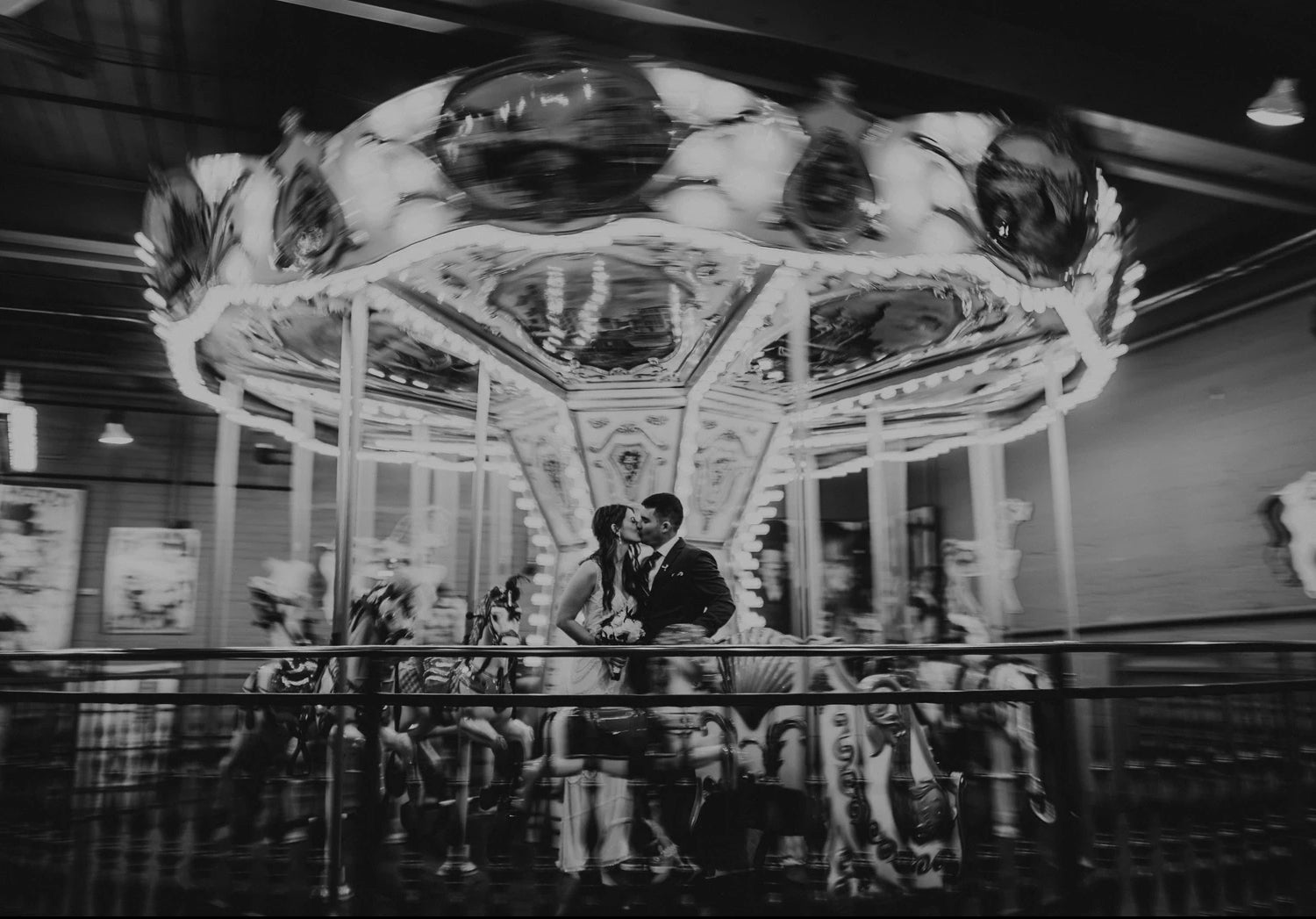 Black and white unique wedding photo of the carnival carousel in motion as the couple pose in front of them 