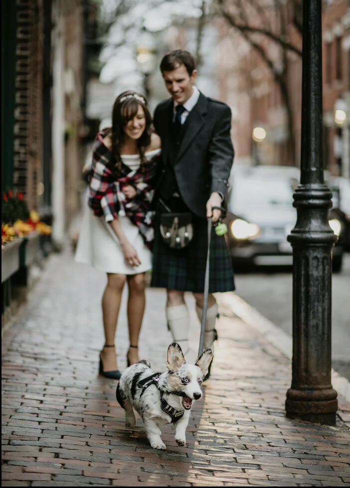 man wearing a kilt and woman wearing a dress with a checkered plaid walking a corgie