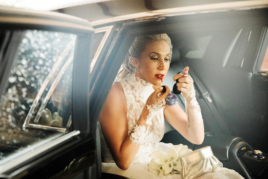 The bride with a short veil over her face is sitting a car, putting on her red lipstick with a compact mirror.