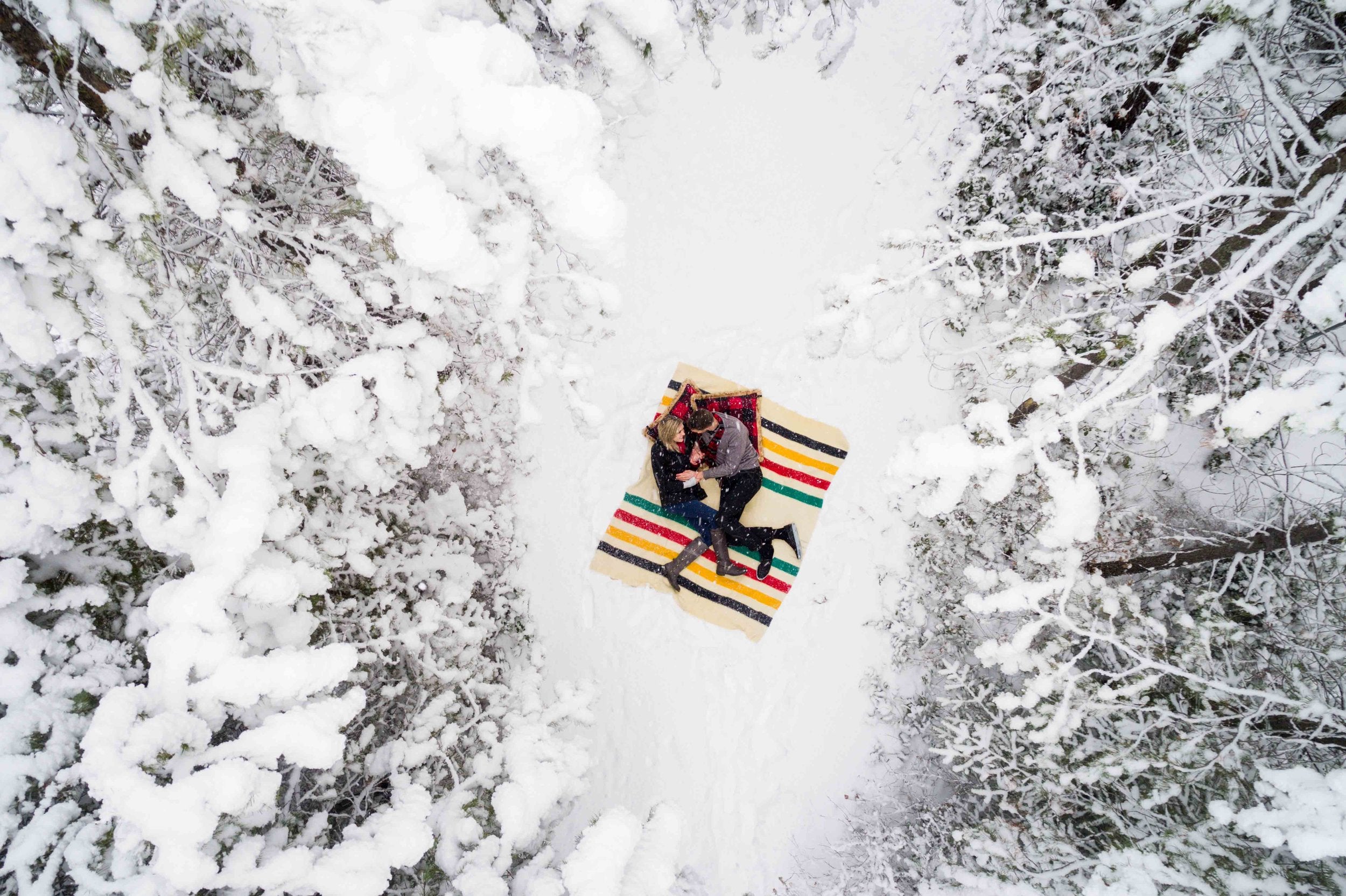 A bird eye’s view of a couple lying on a blanket placed amidst a snow covered forest