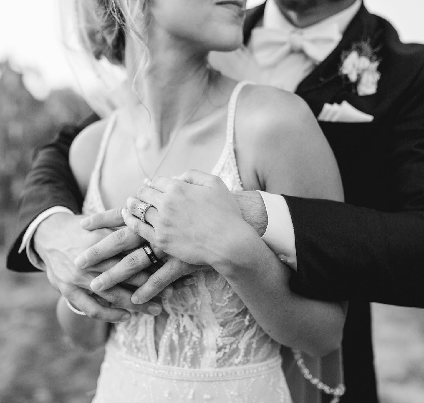 Black and white portrait of a couple showing their rings as the groom hugs the bride from the back and the bride holds his hand