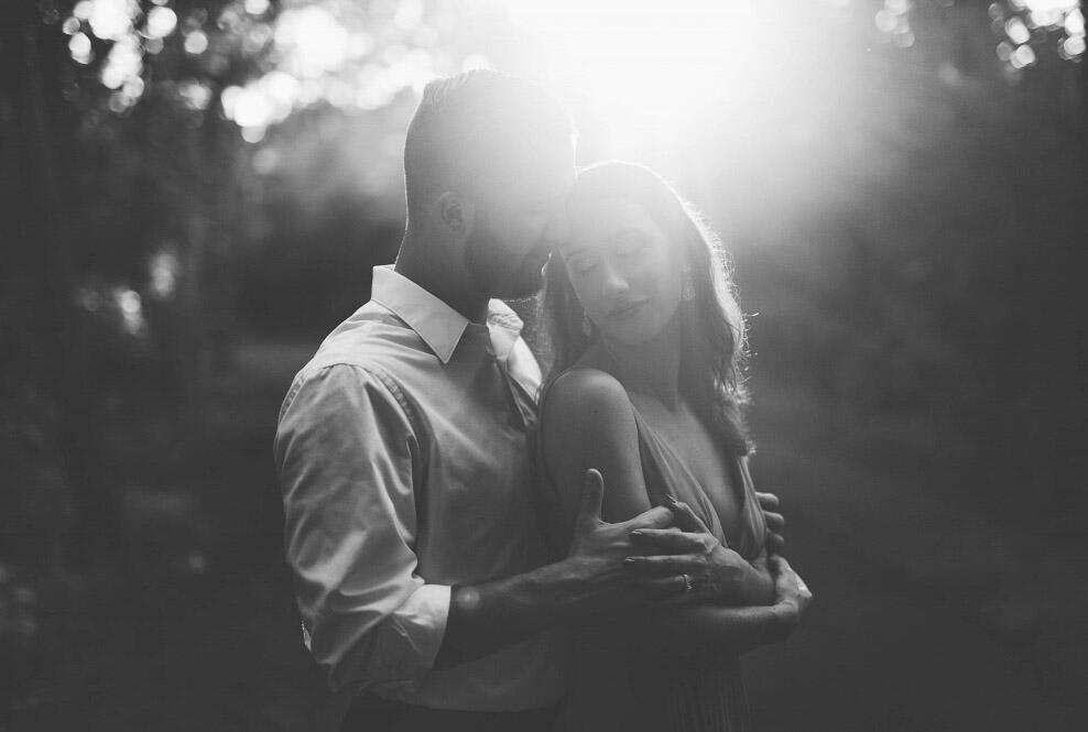 a black and white image of a groom embracing the bride while the sun shine behinds them