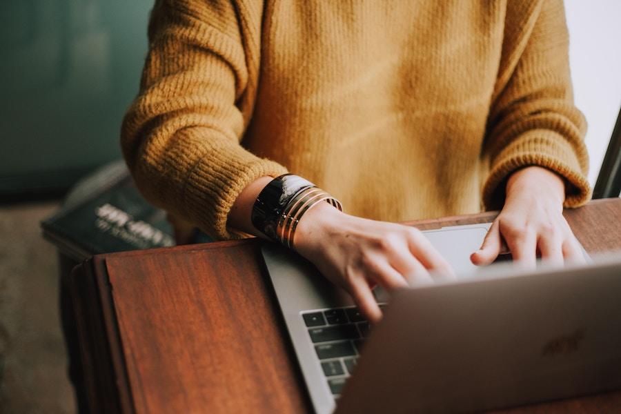 An image of a woman's mustard yellow sweater and her hands as she types on a laptop in front of her, with the background out of focus.