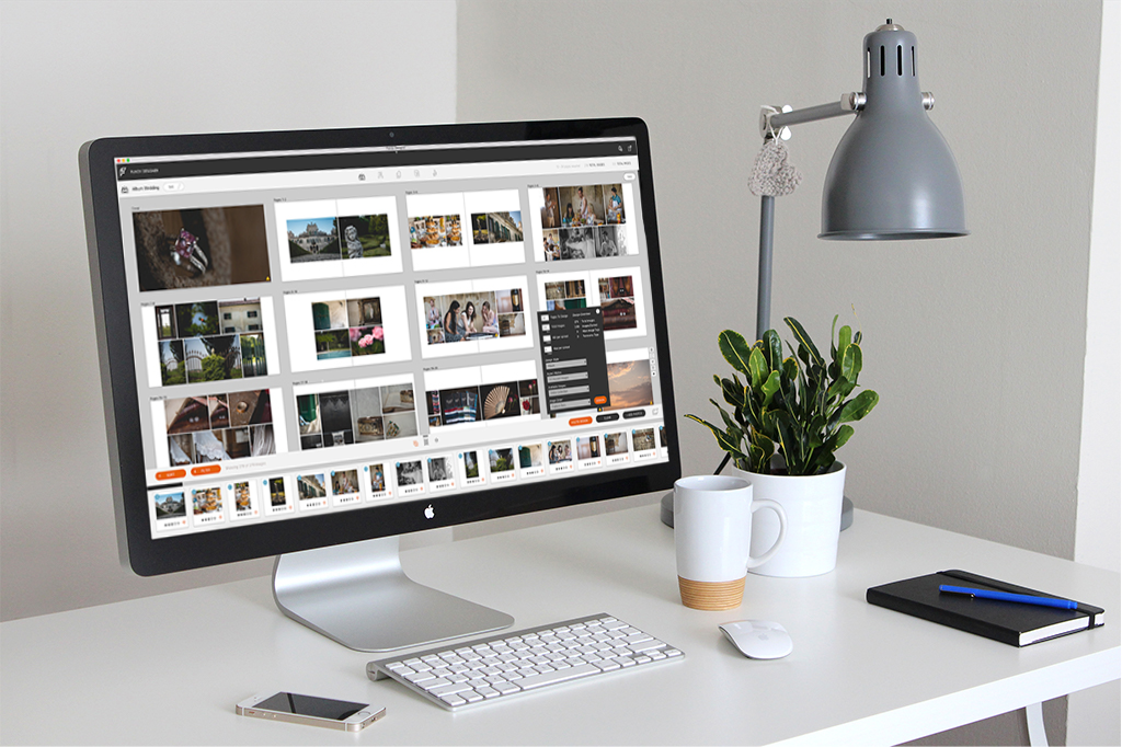 Mac desktop on a desk along with an iPhone, a diary, a table lamp, a coffee mug, and a table plant 