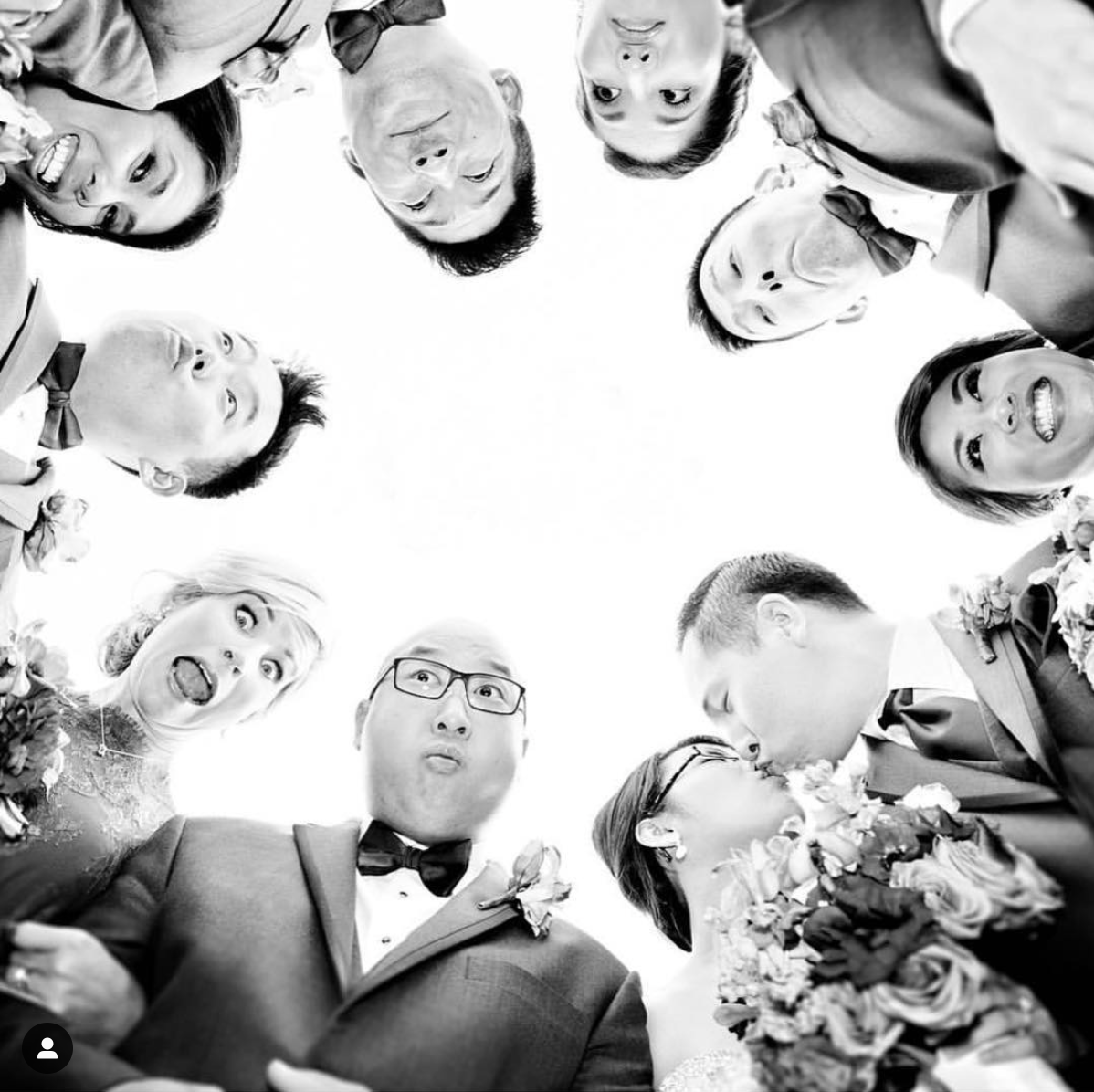 The bridesmaids and groomsmen standing in a circle along with the bride and groom looking down at the camera