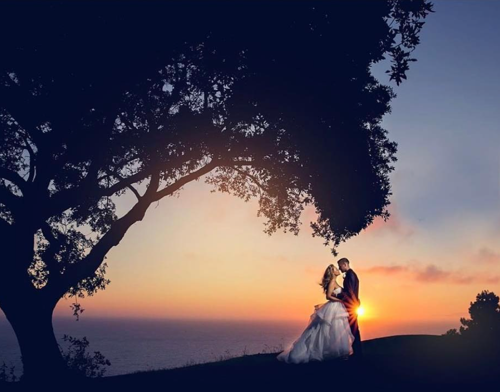 A bride and groom holding each other beside a tree as the sunlight peek in from the groom's side 