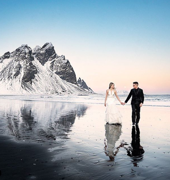 man and woman holding hands walking across a wet surface with a snow covered mountain in the background