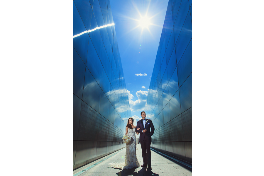 An image of the couple standing side by side, arms interlocked, in between a blue building, with the bride holding her bouquet and the sun shining on them.